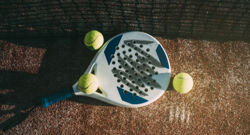 white-and-blue-padel-racket-on-a-brown-court-surrounded-by-3-padel-balls